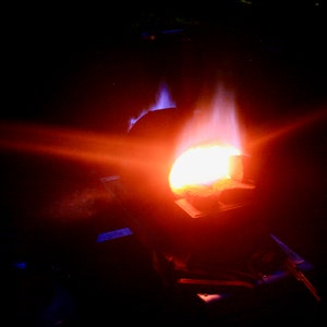 May include: A close-up of molten metal being poured into a mold. The metal is glowing orange and red, and there are flames coming from the mold.