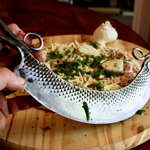 May include: A silver, hand-forged pizza cutter with a decorative handle and a textured blade. The cutter is on a wooden cutting board with a pizza in the background.