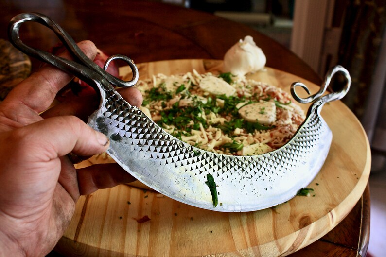 May include: A silver, hand-forged pizza cutter with a decorative handle and a textured blade. The cutter is on a wooden cutting board with a pizza in the background.