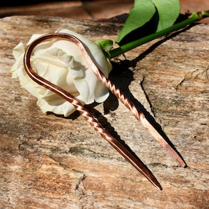 May include: A pair of copper hair pins with a twisted design. The pins are laying on a wooden surface with a white rose.