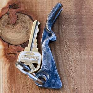 May include: A silver key chain with a key and a handmade bottle opener. The bottle opener is made of dark metal and has a unique shape.