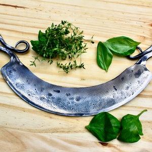 May include: A curved, silver-colored, hand-forged knife with a textured blade and twisted handles. The knife is on a wooden cutting board with fresh herbs.