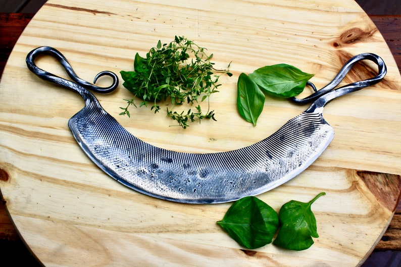 May include: A curved, silver-colored, hand-forged knife with a textured blade and twisted handles. The knife is on a wooden cutting board with fresh herbs.