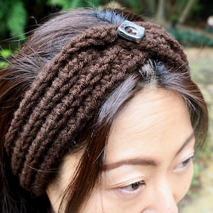 May include: A brown knitted headband with a decorative metal button. The headband is worn by a person with brown hair.