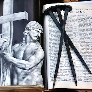May include: An open book with text, featuring a statue of a man holding a cross, and several large nails. The statue is in black and white, and the nails are dark. The book is open to a page of text.