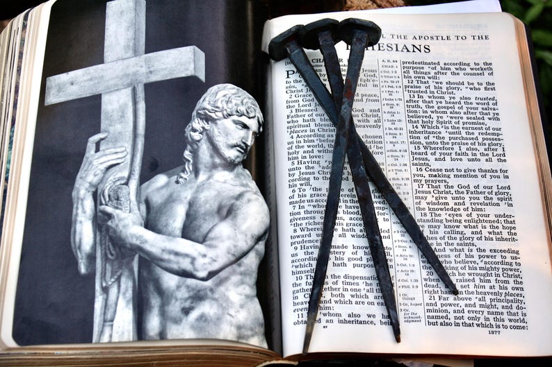 May include: An open book with text, featuring a statue of a man holding a cross, and several large nails. The statue is in black and white, and the nails are dark. The book is open to a page of text.