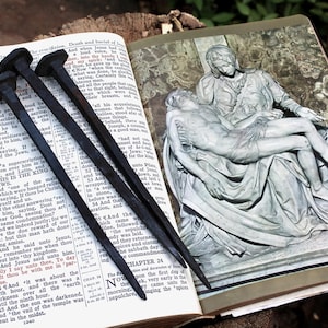 May include: Three dark, aged nails rest on an open Bible, with text from the Book of Luke visible. An illustration of a religious scene is on the adjacent page. The nails are likely symbolic.