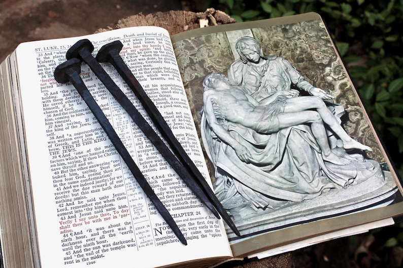 May include: Three dark, aged nails rest on an open Bible, with text from the Book of Luke visible. An illustration of a religious scene is on the adjacent page. The nails are likely symbolic.