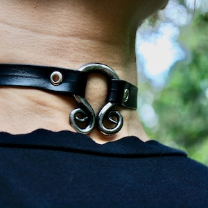 May include: A black leather choker with a silver metal clasp in the shape of a stylized swirl. The choker is worn on a person's neck.