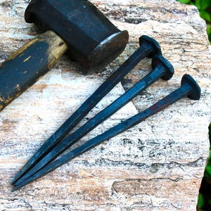 May include: A black metal hammer and three large, dark nails rest on a textured, light-colored stone surface. The hammer has a wooden handle and a dark, rectangular head. The nails are long and tapered, with flat, round heads.