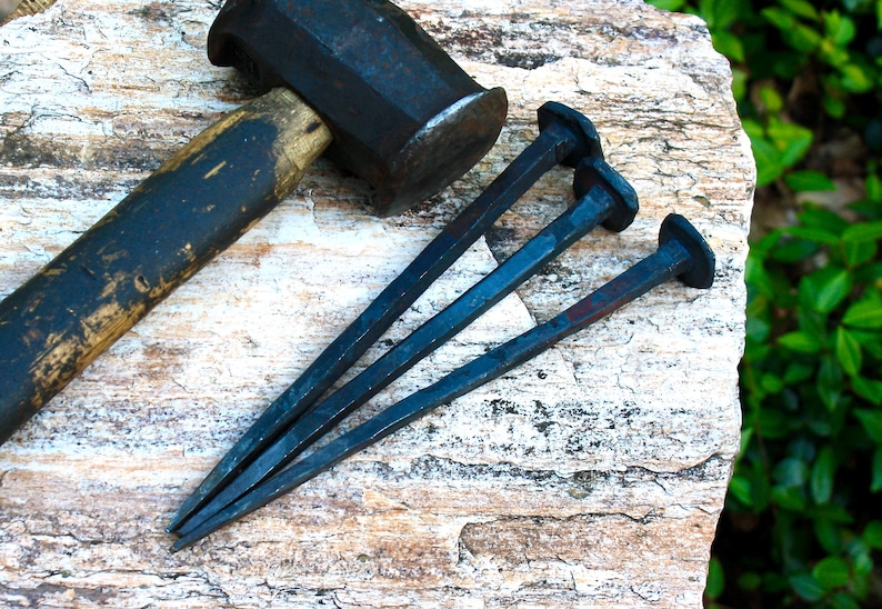 May include: A black metal hammer and three large, dark nails rest on a textured, light-colored stone surface. The hammer has a wooden handle and a dark, rectangular head. The nails are long and tapered, with flat, round heads.