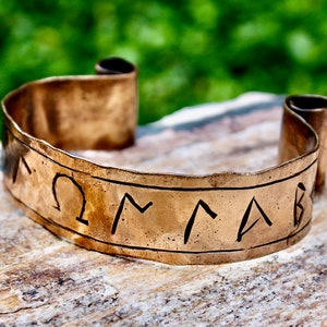 May include: A copper-coloured cuff bracelet with etched symbols. The bracelet is resting on a stone surface with a blurred green background. The ends of the bracelet are rolled up. The etched symbols are dark in colour.