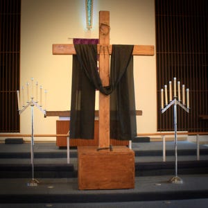 May include: A large wooden cross with a crown of thorns and draped with black fabric stands in a church. Two silver candelabras with lit candles flank the cross. The cross is on a wooden base.