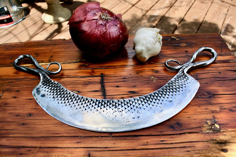 May include: A hand-forged, crescent-shaped cheese knife with a textured blade and twisted handles. The knife is resting on a wooden surface with a red onion and a garlic clove beside it.