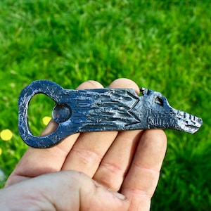 May include: A hand-forged, black metal bottle opener. The opener features a stylized boar head design with a star-shaped element and a circular opening for the bottle cap. The opener is held against a backdrop of green grass.