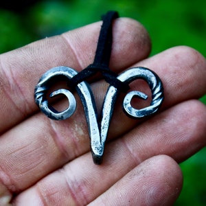 May include: A silver metal pendant shaped like a ram's head with curled horns. The pendant is hanging from a black cord.