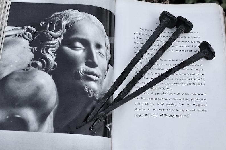 May include: Three dark, square-headed nails rest on an open book featuring a black and white image of a sculpture. The nails are positioned near the text, adding a rustic, industrial contrast to the classical art.