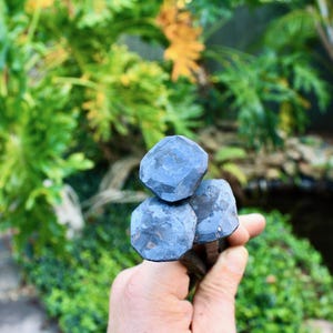 May include: Three dark blue, faceted metal objects are held in a hand, with a blurred green background of foliage. The objects have a geometric, almost gem-like appearance. The overall aesthetic suggests a handcrafted or artisan product.