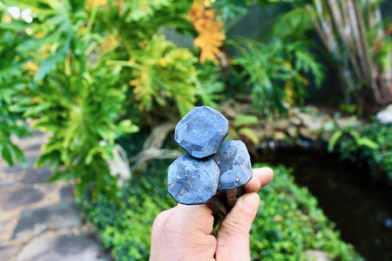 May include: Three dark blue, faceted metal objects are held in a hand, with a blurred green background of foliage. The objects have a geometric, almost gem-like appearance. The overall aesthetic suggests a handcrafted or artisan product.