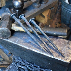 May include: Close-up of blacksmith tools, including three large, hand-forged nails with square heads, resting on an anvil. A hammer and other tools are visible in the background, suggesting a workshop setting. The nails are made of dark metal.