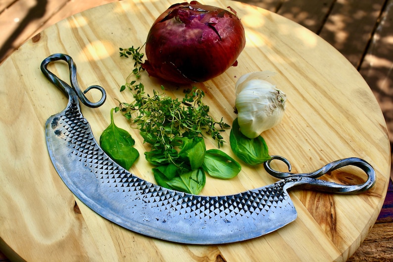 May include: A curved, hand-forged, steel knife with a textured blade and twisted handles sits on a wooden cutting board with a red onion, garlic, thyme, and basil.