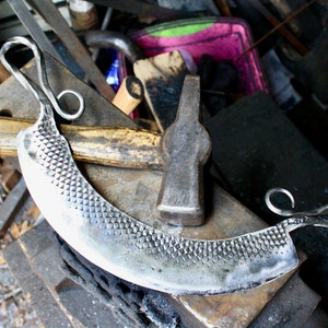 May include: A hand-forged, silver-colored, curved cheese knife with a textured blade and two looped handles. The knife is resting on a metal anvil with a wooden surface. A hammer with a wooden handle is lying on the anvil.