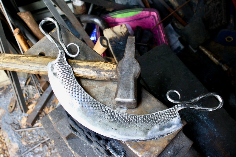 May include: A hand-forged, silver-colored, curved cheese knife with a textured blade and two looped handles. The knife is resting on a metal anvil with a wooden surface. A hammer with a wooden handle is lying on the anvil.