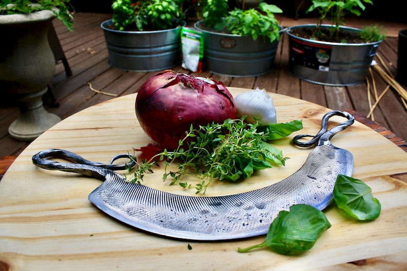 May include: A close-up of a  handmade, wavy,  metal  grater with a wooden handle. The grater is on a wooden cutting board with a red onion, garlic, and fresh herbs.