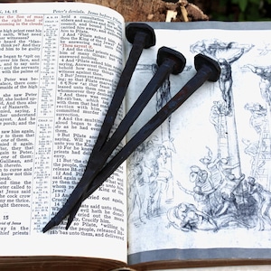 May include: An open book with text and illustrations, featuring three dark, iron nails. The nails are positioned over a drawing of a crucifixion scene. The book's pages are aged, suggesting a historical or religious theme.