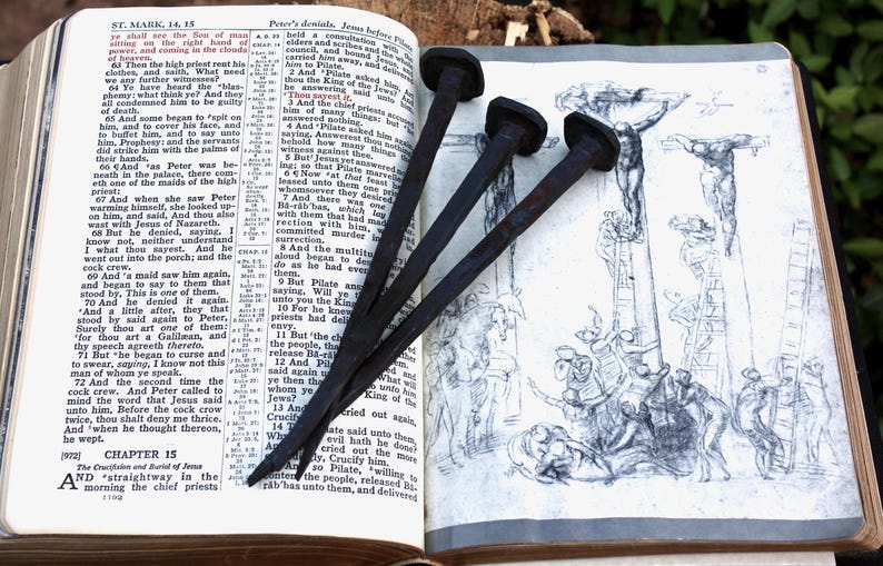 May include: An open book with text and illustrations, featuring three dark, iron nails. The nails are positioned over a drawing of a crucifixion scene. The book's pages are aged, suggesting a historical or religious theme.