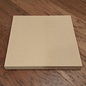 May include: A square, light brown wooden board, possibly for crafts or DIY projects. The board is resting on a dark brown wooden floor with visible grain patterns. The board appears to be made of pressed wood or similar material.