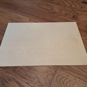 May include: A rectangular, light-colored wooden board with visible wood grain. The board is positioned on a dark brown wooden surface, creating a contrast in tones. The wood grain on the board is subtle and natural.