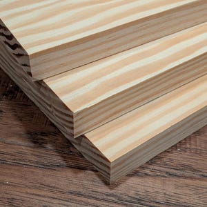 May include: Three stacked rectangular wooden boards featuring a light tan color with prominent, wavy, darker brown grain patterns running lengthwise. The edges of the boards are straight and smooth, showcasing the layered texture of the wood grain.