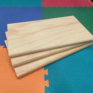 May include: A stack of three unfinished wooden boards with rounded edges. The boards are light-colored with visible wood grain. The boards are stacked on a colorful foam play mat.