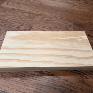 May include: A stack of unfinished, light-colored wood panels. The wood grain is visible, with a mix of light and medium brown tones. The panels are rectangular and appear to be ready for use in a woodworking project.