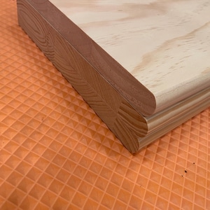 May include: Close-up of a wooden board with a light-colored surface and a darker, reddish-brown edge. The edge has a decorative, curved design. The board is likely for woodworking or crafting projects.