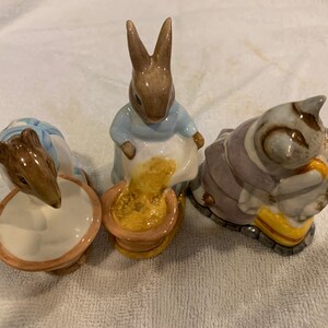 May include: Three ceramic figurines of Beatrix Potter characters: Peter Rabbit, a mouse, and a cat. Peter Rabbit is pouring a yellow substance from a bag into a brown pot. The mouse is dipping its nose into a white bowl. The cat is holding a smaller cat in its arms.