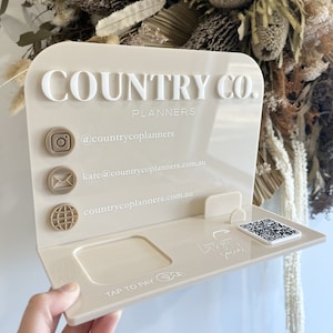 May include: A beige acrylic sign with the text "COUNTRY CO. PLANNERS" in white lettering. The sign has icons for Instagram, email, and a website, along with contact information. The sign also has a space for a card reader and a QR code.