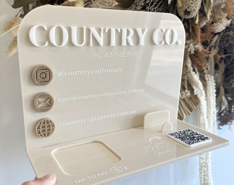May include: A beige acrylic sign with the text "COUNTRY CO. PLANNERS" in white lettering. The sign has icons for Instagram, email, and a website, along with contact information. The sign also has a space for a card reader and a QR code.