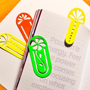 May include: A collection of colorful, die-cut metal bookmarks in yellow, orange, and green, placed in an open notebook. The bookmarks feature floral and geometric designs. The notebook pages have printed text.