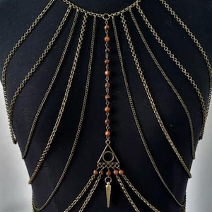Peut inclure: Un collier de corps de couleur bronze avec plusieurs rangées de chaînes fines. Le collier présente un pendentif central avec un triangle et une pointe pendante, orné de petites perles marron. Le bijou est présenté sur un mannequin noir.