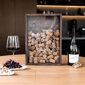 Puede incluir: Una caja expositora rectangular de madera oscura llena de corchos de vino, con un dibujo lineal blanco de un mapa mundial, un avión y el texto "travel far travel wide" con la fecha "01.08.2024" impresa en el panel frontal de cristal.