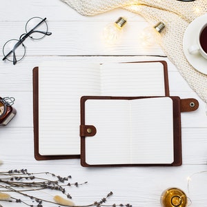 May include: Two open brown leather journals with lined pages on a white wooden surface. The journals are surrounded by decorative elements, including string lights, a knitted blanket, and a small jar of honey.