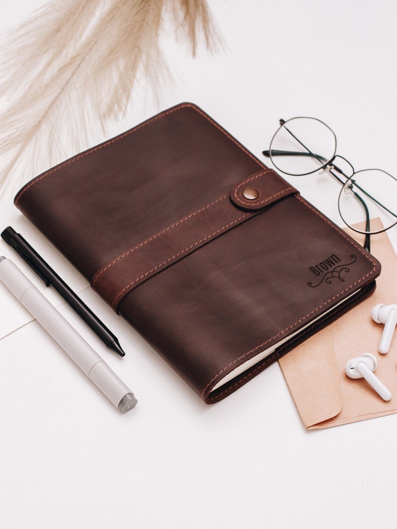 May include: A brown leather journal with a button closure and the word "Brown" engraved on the cover. The journal is lying on a white surface with a pen, glasses, and earbuds nearby.