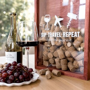 Puede incluir: Una caja de madera con frente de cristal llena de corchos de vino, con iconos blancos de una copa, un avión y un sacacorchos junto al texto "SIP TRAVEL REPEAT Collect the good stuff". Al lado hay una botella de vino, una copa de vino tinto y uvas rojas.