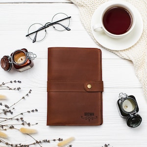 May include: A brown leather journal with a snap closure and the word "BROWN" embossed on the front. The journal is on a white wooden surface with a cup of tea, glasses, and candles.