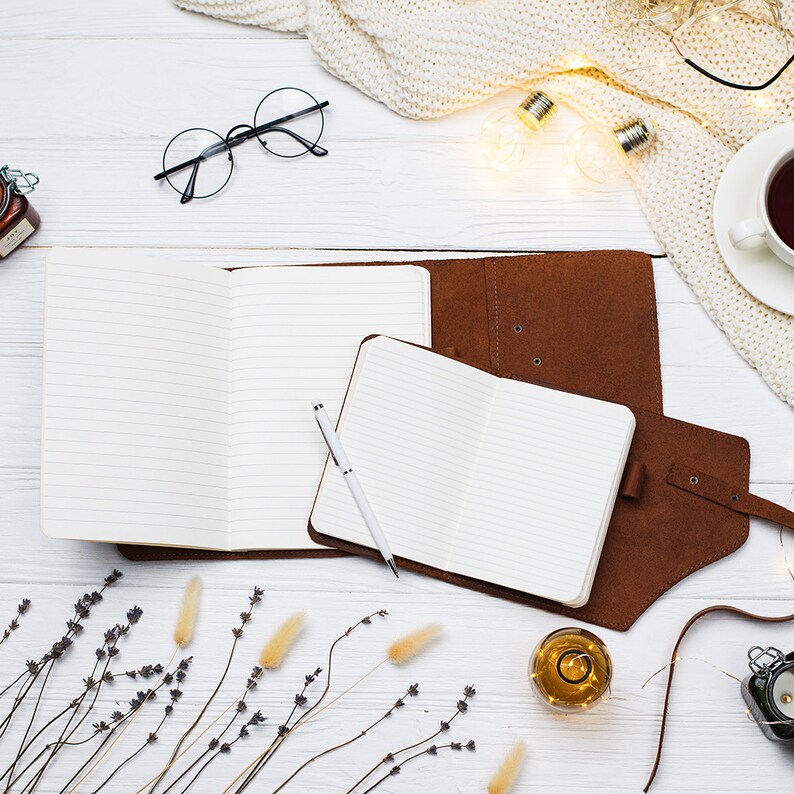 May include: A flat lay of a white wooden surface with two open notebooks, a pen, a jar of honey, a cup of tea, a knitted blanket, string lights, dried lavender, and a small glass bottle with a cork.
