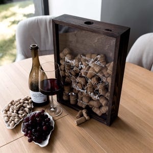 Puede incluir: Una caja expositora de madera oscura llena de corchos de vino, con un mapa mundial impreso en blanco, un icono de avión, el texto "Travel far, Travel wide" y la fecha "01.08.2024". Al lado hay una botella de vino, una copa y platos blancos.
