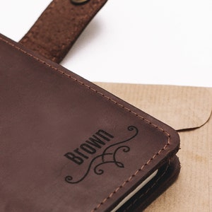 May include: A brown leather journal with a stitched edge and a brass button closure. The journal has the word "BROWN" engraved on the front cover with a decorative flourish.