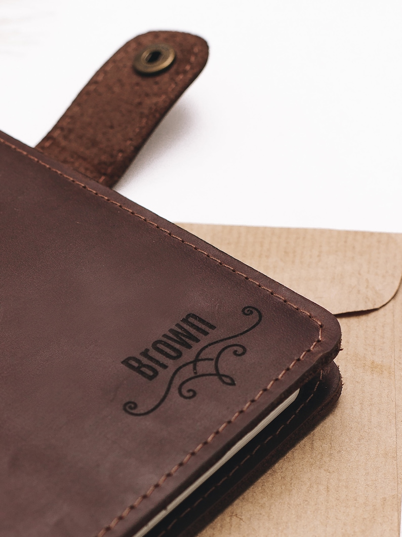 May include: A brown leather journal with a stitched edge and a brass button closure. The journal has the word "BROWN" engraved on the front cover with a decorative flourish.